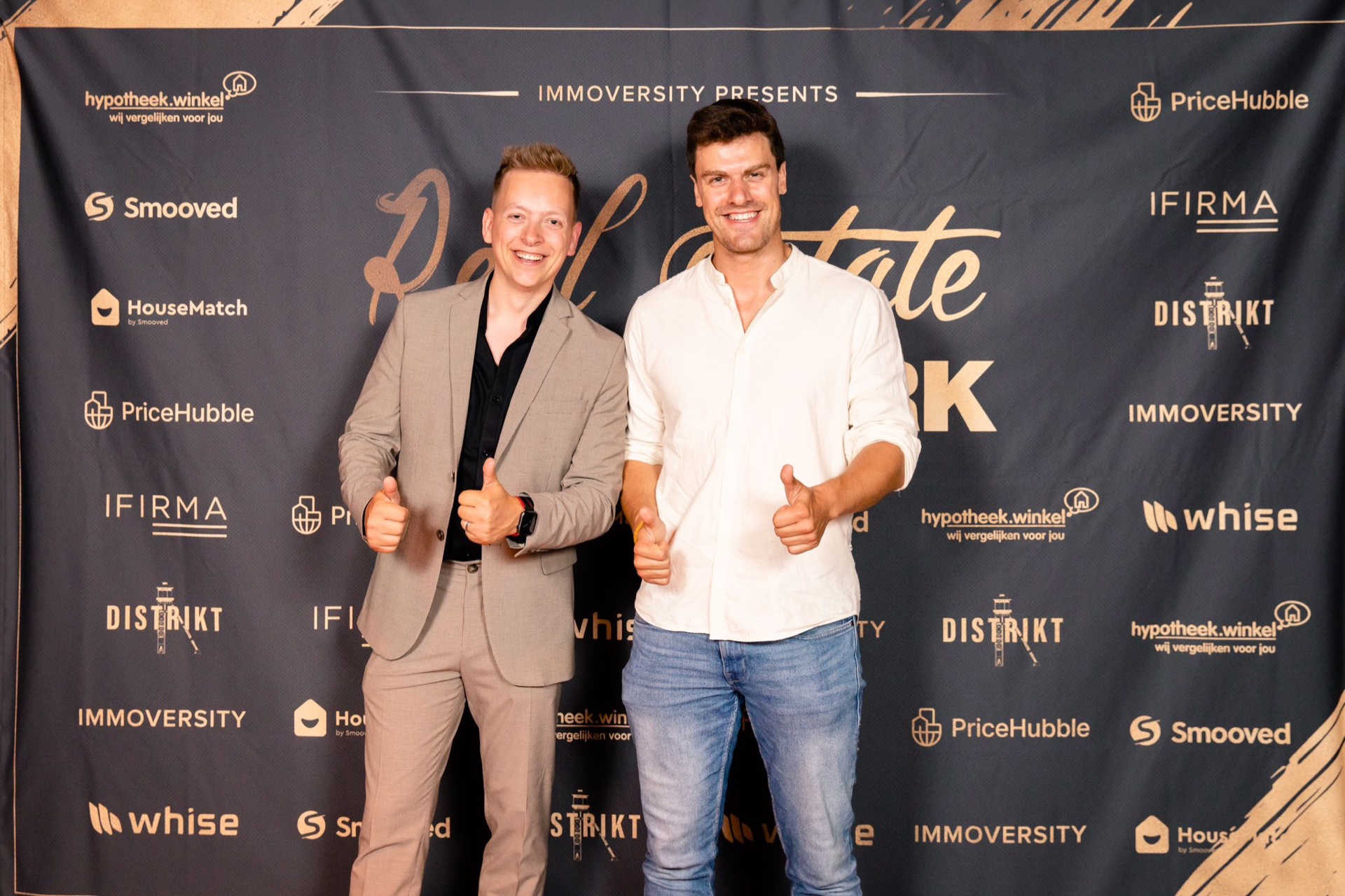 Twee mensen met de duim omhoog voor de perswand van het Immoversity-event.
